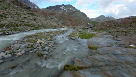 Freestyle-drone-flight-over-bridge-and-water-torrent-stream-flowing-at-Fellaria-glacier-in-Valmalenco-of-Valtellina,-Italy