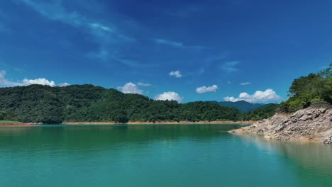 Low-altitude-flight-over-natural-lake-named-Feicui-Reservoir-with-green-jungle-hills-,-sandy-shore-and-blue-sky