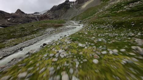 Racing-fpv-drone-flying-at-high-speed-over-water-stream-flowing-at-Fellaria-glacier-in-Valmalenco-of-Valtellina,-Italy