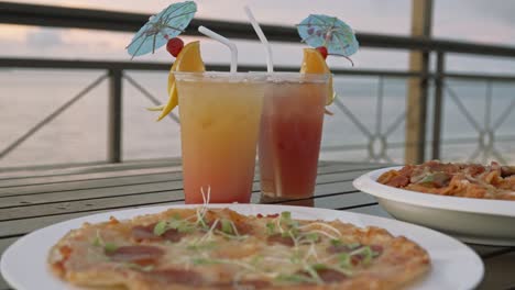 Pizza,-Pasta-Und-Cocktailgetränke-Auf-Einem-Tisch-Am-Strand-Bei-Einem-Friedlichen-Sonnenuntergang