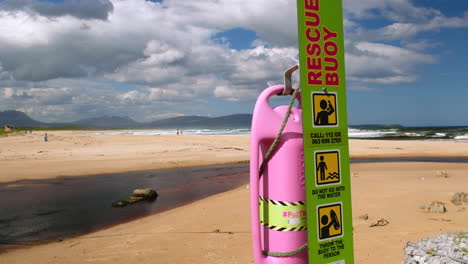 Pink-rescue-buoy-at-Kleinmond's-blue-flag-beach,-nice-day-next-to-lagoon