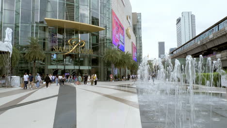 A-water-fountain-in-front-of-Siam-Paragon-and-the-Siam-BTS-Station-with-a-train-waiting-and-people-walking-about,-Bangkok,-Thailand