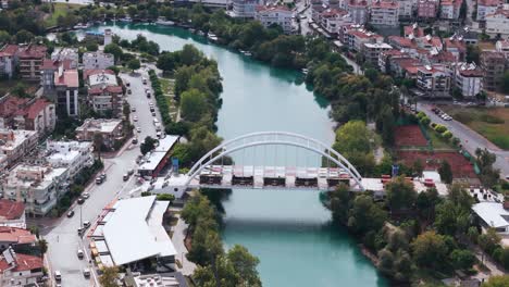 Aerial-view-orbiting-archway-bridge-over-the-curving-Manavgat-river-in-the-city-centre-of-the-Antalya-region,-Turkey