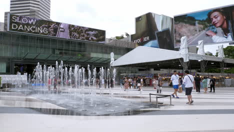 A-water-fountain-in-front-of-Siam-Paragon-and-the-Siam-BTS-Station-with-a-train-waiting-and-people-walking-about,-Bangkok,-Thailand