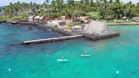 Close-up-panning-aerial-shot-tracking-two-stand-up-paddleboarders-in-front-of-the-historic-Kamakahonu-House-in-Kailua-Kona-on-the-Big-Island-of-Hawai'i