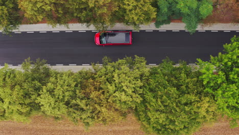 Drone-top-down-asymmetrical-follow-shot-of-red-minivan-camper-with-black-roof-with-solar-panels-driving-along-a-narrow-unmarked-road-in-alley-of-trees