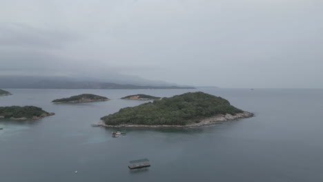 Drone-shot-flying-over-the-water-to-the-small-islands-in-front-of-Ksamil-Albania-in-the-morning-on-a-cloudy-grey-day-with-boats-around-and-haze-in-the-sky-LOG