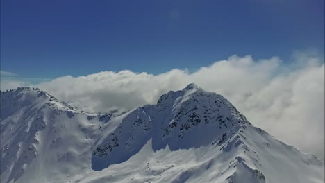 Luftaufnahme-Eines-Schneebedeckten-Berggipfels-Mit-Wolken,-Die-über-Den-Gipfeln-Vor-Blauem-Himmel-Hängen