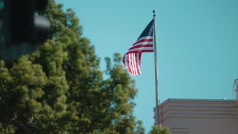 A-Shot-of-an-American-Flag-Flapping-on-Top-of-a-Building-in-the-Wind-with-a-Tree-and-a-Stoplight-in-the-Foreground-at-Daytime