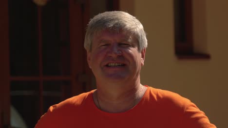 Grey-haired-man-in-orange-t-shirt-smiling-friendly-toward-camera-close-up-portrait