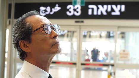 A-Japanese-businessman-stands-at-Ben-Gurion-Airport-in-Israel,-watching-the-arrivals-and-departures-board,-with-an-exit-sign-in-Hebrew-in-the-background