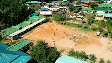 4K-Cinematic-sport-aerial-drone-footage-of-children-playing-on-a-footbal-field-in-the-mountain-village-of-Doi-Pui-next-to-Chiang-Mai,-Thailand-on-a-sunny-day