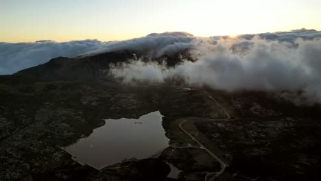 From-an-aerial-vantage-point,-behold-a-panoramic-view-of-Portugal's-highest-peak-in-the-Serra-de-Estrella,-where-dense-cloud-formations-share-the-sky-with-a-backdrop-of-a-rising-sunset