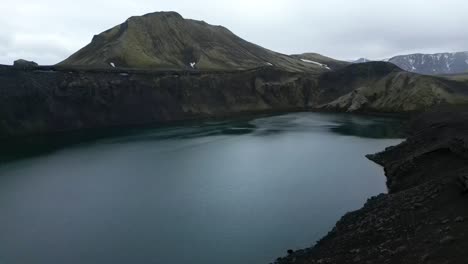 Vista-Aérea-Desde-El-Lago-Del-Cráter-Blahylur-En-Las-Tierras-Altas-De-Islandia-Durante-El-Verano,-Rodeado-De-Formas-Verdes-Y-Grises,-Hermosas-Montañas-Y-Caminos-Oscuros