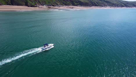An-aerial-drone-follows-a-luxury-speed-boat-along-the-Cornish-coastline,-traversing-turquoise-waters