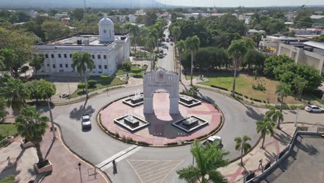Aerial-backwards-shot-of-Triumphal-Arch-and-driving-vehicle-in-roundabout-during-sunny-day---San-Juan,-Dominican-Republic