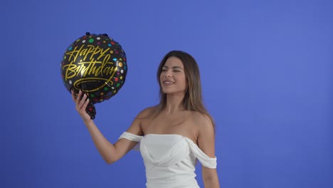 A-young-woman-dances-with-a-Happy-Birthday-balloon,-isolated-on-a-blue-studio-background
