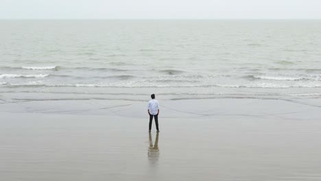 Reversing-Drone-Shot-of-a-Man-Standing-Alone-by-the-Sea-and-Staring-Sadly-or-Melancholicly-at-the-Grey-Horizon-on-a-Cloudy-Day,-Kuakata-Sea,-Bangladesh