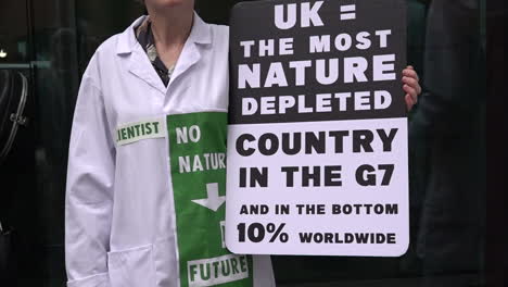 In-slow-motion-a-scientist-in-a-white-coat-holds-a-placard-that-reads,-“UK-=-the-most-nature-depleted-country-in-the-G7-and-in-the-bottom-10%-worldwide”