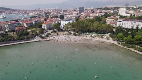 Aerial:-Croatia,-Bacvice-Beach,-Split,-with-swimmers-and-cityscape-backdrop