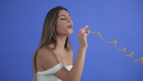Beautiful-girl-blows-a-yellow-party-streamer,-isolated-on-blue-background