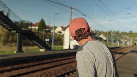 Young-man-wearing-hat-and-standing-on-railway-platform-watching-for-train-coming