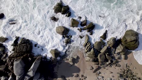 Drohne-Stößt-Gegen-Felsen-Am-Strand,-Während-Wellen-über-Sie-Krachen