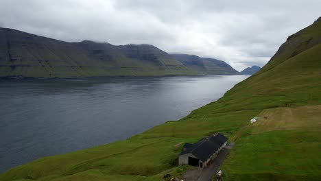 Flyover-towards-a-calm-fjord-from-Mikladalur-and-striking-volcanic-landscape-of-Kunoy,-Faroe-Islands