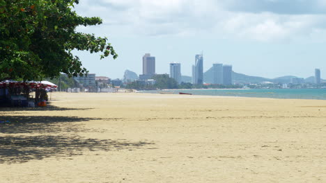 Pattaya-beachfront-with-big-umbrellas-on-the-left-then-the-beautiful-clean-sand,-shoreline-with-boats-and-waves-rolling,-in-the-horizon-is-the-city-revealing-hotels,-condos,-and-the-mountains