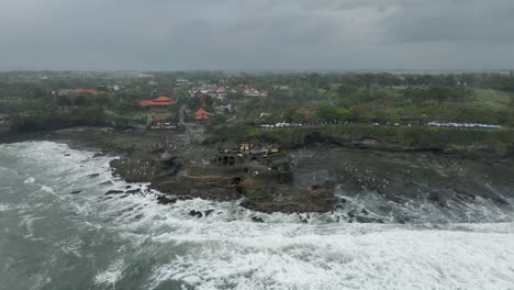 Grey-overcast-aerial-orbits-Tanah-Lot-temple-as-waves-crash-on-shore
