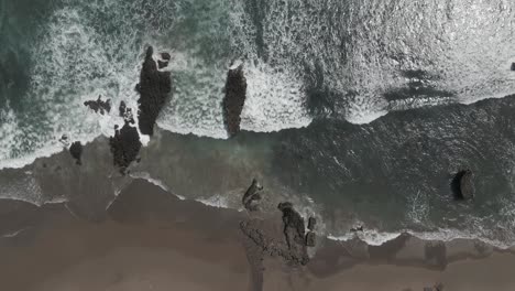 Aerial-looks-directly-down-onto-ocean-waves-on-rocks-near-sand-beach