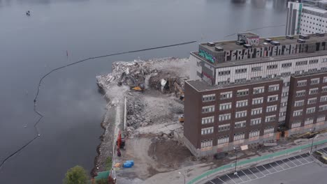 Drone-shot-orbiting-building-revealing-building-with-three-diggers-working-on-removing-rubble-by-the-water-in-Liljeholmsviken,-Liljeholmen,-Stockholm,-Sweden