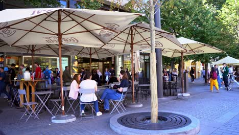 Panorama-of-people-sitting-talking-in-a-restaurant-with-giant-umbrellas-in-the-middle-of-streets-with-tourists-constantly-passing-by-on-a-sunny-day-in-Prague,-Czech-Republic