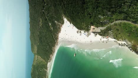 Vertical-drone-shot-of-sandy-beach-with-clear-ocean-and-green-hills-in-Brazil,-South-America---Birds-eye-shot
