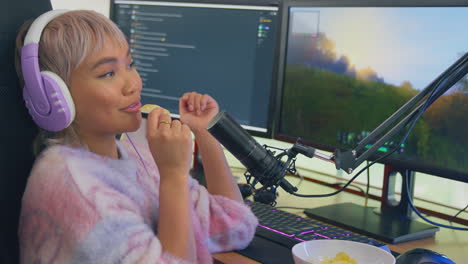 Woman-Eating-Snacks-Whilst-Gaming-At-Home-Sitting-At-Desk-Wearing-Headphones-For-Live-Stream