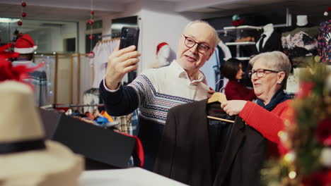 Senior-husband-and-wife-in-festive-ornate-fashion-shop-talking-with-grandson-over-online-videocall,-asking-him-to-pick-between-stylish-blazers-to-buy-as-gift-before-xmas-family-event