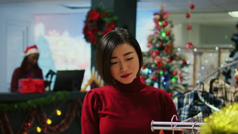 Portrait-of-happy-asian-woman-in-red-sweater-browsing-through-clothes-racks-in-festive-shopping-mall-clothing-store-during-Christmas-holiday-season.-Shopper-looking-to-buy-gifts-in-fashion-boutique