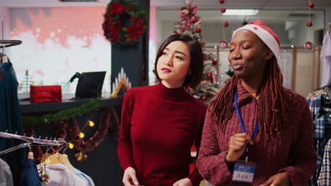 Tracking-shot-of-helpful-retail-assistant-in-festive-decorated-clothing-store-showing-asian-customer-beautiful-red-garments,-ready-to-be-worn-at-Christmas-themed-office-party