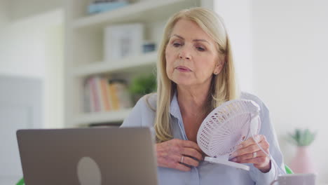 Menopausal-Mature-Woman-Having-Hot-Flush-At-Home-Cooling-Herself-With-Fan-Connected-To-Laptop