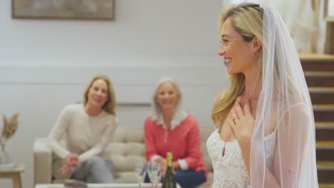 Grandmother-With-Mother-Watching-Adult-Daughter-Choosing-And-Trying-On-Wedding-Dress-In-Bridal-Store