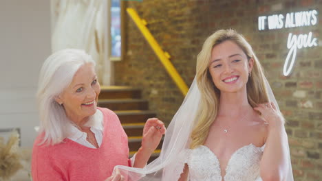 Grandmother-Watching-Adult-Daughter-Choosing-And-Trying-On-Wedding-Dress-In-Bridal-Store