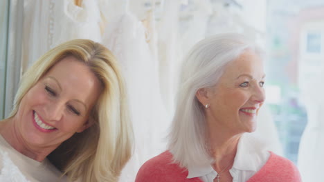 Grandmother-With-Adult-Granddaughter-Watching-Mother-Choosing-Wedding-Dress-In-Bridal-Store
