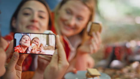 Woman-Taking-Video-Of-Friends-On-Camping-Holiday-In-Forest-Lying-In-Tent-Eating-S'mores
