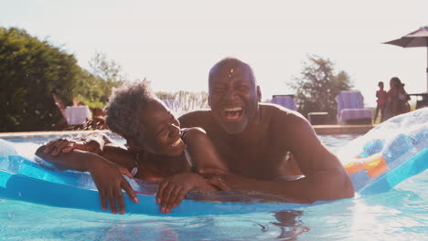 Portrait-Of-Smiling-Senior-Couple-On-Summer-Holiday-Relaxing-In-Swimming-Pool-On-Inflatable-Airbed