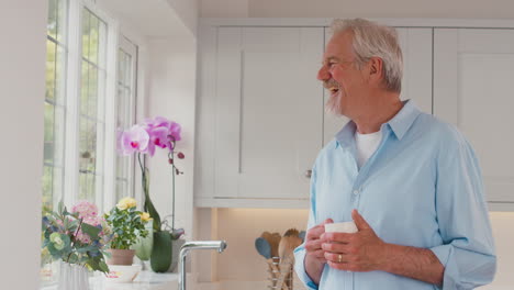 Retired-Senior-Man-Standing-By-Window-In-Kitchen-At-Home-Drinking-Coffee