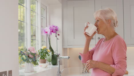 Retired-Senior-Woman-Standing-By-Window-In-Kitchen-At-Home-Drinking-Coffee