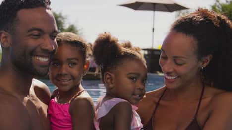 Family-On-Summer-Holiday-With-Two-Girls-Being-Held-In-Swimming-Pool-By-Parents-And-Splashing