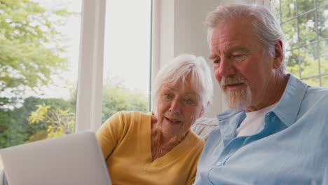 Retired-Senior-Couple-Sitting-On-Sofa-At-Home-Using-Laptop-Online-Shopping-Or-Booking-Holiday