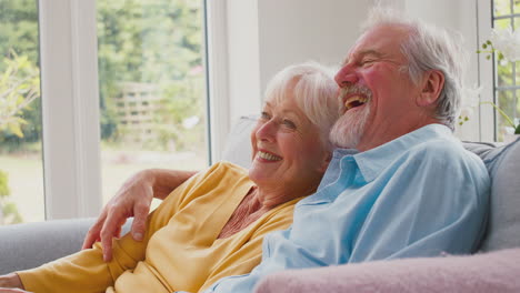 Retired-Senior-Couple-With-Remote-Control-Sitting-On-Sofa-At-Home-Watching-TV-And-Laughing-Together
