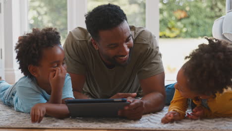 Vater-Zu-Hause-Mit-Kindern,-Die-Auf-Dem-Teppich-Liegen-Und-Mit-Einem-Digitalen-Tablet-Spielen